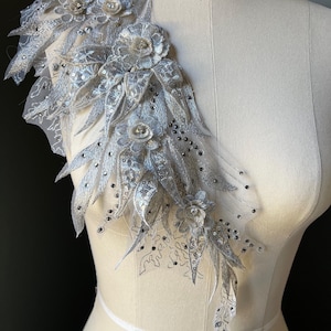 May include: A silver lace applique with floral and leaf designs, embellished with sequins and small beads. The applique is displayed on a mannequin, showcasing its intricate details and delicate texture. Ideal for bridal or formal wear.