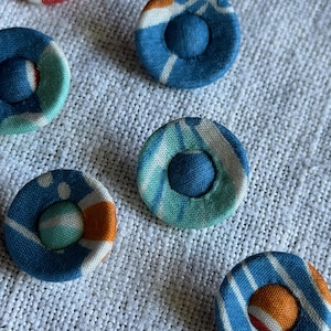 May include: A collection of circular fabric-covered buttons featuring various patterns in shades of blue, orange, red, and white. Each button has a central raised fabric-covered dome surrounded by a flat, circular rim with distinct textile prints.