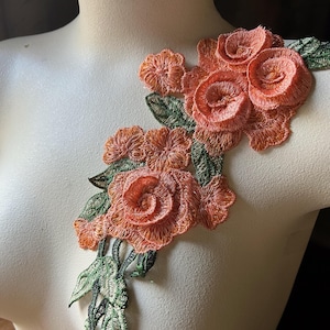 Puede incluir: Aplicación floral bordada en naranja con hojas y tallos verdes. La aplicación presenta flores detalladas en forma de rosa y se muestra en un maniquí. El bordado tiene un aspecto texturizado.
