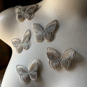 May include: Five silver glitter butterfly appliques on a white background. The butterflies are made of sheer fabric with a delicate embroidered design.
