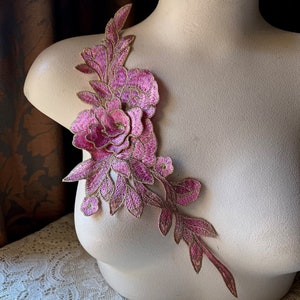 Könnte beinhalten: Eine rosa und goldene Blumenapplikation mit einer großen Rose und kleineren Blumen. Die Applikation befindet sich auf einem weißen Hintergrund.