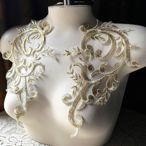 May include: Gold embroidered lace applique displayed on a mannequin. The applique features intricate floral and scroll designs. The delicate lace is a light gold color, with a sheer backing. The mannequin is a neutral color, and the background is dark.