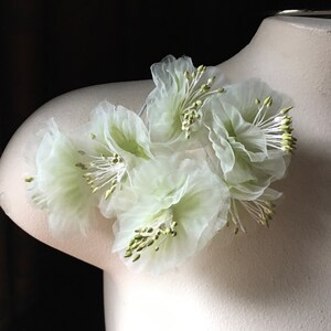 May include: A cluster of delicate, white, sheer fabric flowers with green centers. The flowers are arranged in a circular shape and are attached to a white surface.