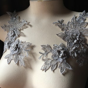 May include: Two silver lace appliques with floral designs. The appliques feature delicate leaves and flowers with a shimmery finish.