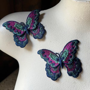 May include: Two embroidered butterfly patches. The butterflies feature a vibrant color palette of magenta, teal, and navy blue. The wings are intricately detailed with a textured appearance. The patches are likely for decorative purposes, such as embellishing clothing or accessories.