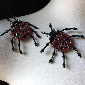 May include: Two black beaded spider brooches with red and black rhinestone accents. The spiders have eight legs and a round body.