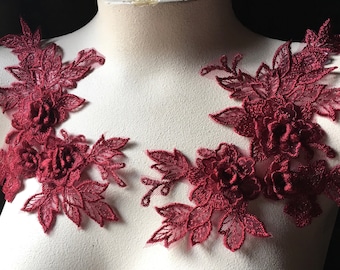 May include: Two burgundy lace appliques with floral designs. Each applique features a cluster of three-dimensional flowers with delicate leaves.