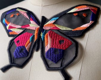 Puede incluir: Aplicación de mariposa colorida con contorno negro y alas transparentes. La mariposa presenta bordados geométricos en tonos rosa, naranja, azul y verde. Ideal para ropa o accesorios.