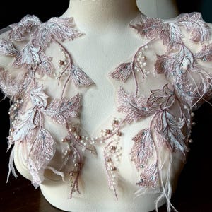 May include: A delicate, blush-colored lace applique featuring embroidered leaf and feather details. The design includes small pearl and crystal accents, creating a refined, elegant aesthetic. The applique is displayed on a mannequin, showcasing its potential for bridal or formal wear.