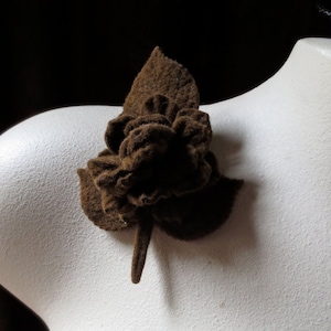 May include: A brown felt flower brooch with a detailed, layered design. The brooch is attached to a white mannequin, showcasing its shape and texture. The flower has a stem and a leaf.