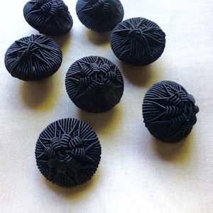 May include: Seven black buttons with a starburst design. The buttons are made of a woven fabric and have a raised, textured surface.
