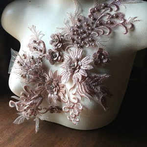 May include: A delicate rose gold lace applique with pearl embellishments and rhinestones. The applique features floral designs and intricate details.