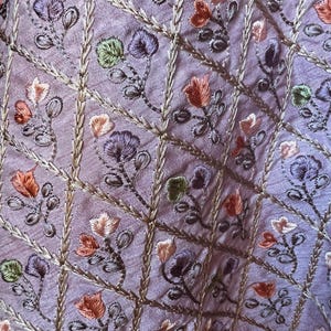 May include: Close-up of a lavender fabric with embroidered floral patterns. The embroidery features flowers in shades of orange, purple, and green, with gold-colored stems and a grid-like design. The fabric has a subtle sheen.