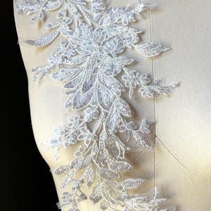 May include: A white floral lace applique with sequins, displayed on a mannequin. The applique features a delicate design of leaves and flowers, with shimmering sequins adding a touch of sparkle. This decorative piece is suitable for bridal wear or other embellishment projects.