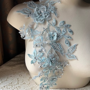 Könnte beinhalten: Eine hellblaue Blumenapplikation mit silbernen Pailletten und Perlen. Die Applikation zeigt zarte Blumen und Blätter, perfekt, um Kleidung oder Bastelarbeiten einen Hauch von Eleganz zu verleihen.