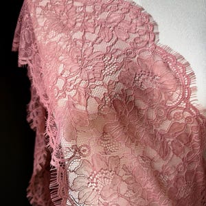 May include: A dusty rose colored lace fabric featuring an intricate floral pattern with scalloped edges. The delicate textile displays a detailed mesh background with woven flower motifs and fine fringe along the borders of the material.