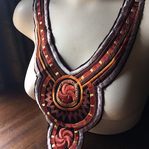 May include: A red, orange, and silver embroidered neckline trim with a geometric design. The trim is on a white mannequin.
