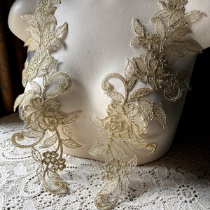 Peut inclure: Deux appliques en dentelle dorée avec des motifs floraux. Les appliques sont sur un mannequin blanc et un fond en dentelle blanche.