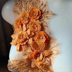 May include: Orange floral lace applique with rhinestone accents. The applique features detailed embroidery of flowers and leaves, with a delicate net backing. The design is embellished with small, sparkling rhinestones, adding a touch of glamour to the craft.