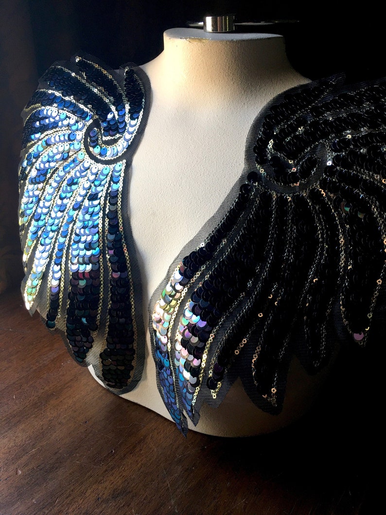 May include: Two iridescent blue and black sequined wings on a white mannequin. The wings are made of sequins and mesh fabric.