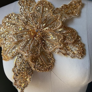 Pode incluir: Aplique floral dourado com lantejoulas, intrincado trabalho de contas e um strass central. O aplique foi concebido para ser cosido em tecido, adicionando um elemento decorativo a vestuário ou acessórios. O design é exibido num manequim.