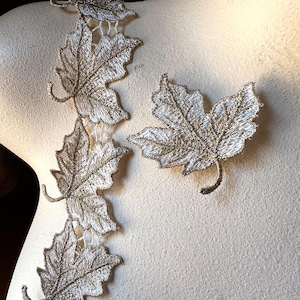 May include: Close-up of embroidered silver maple leaf appliques. The leaves are intricately detailed with delicate stitching and attached to a fine, open-weave backing. One leaf is separate, while others are connected in a line, suggesting a trim or embellishment.