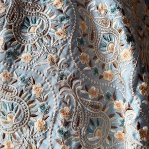 May include: A light blue fabric featuring an intricate, dense embroidery pattern of paisley shapes and small floral motifs. The embroidery uses a colour palette of soft yellow, muted brown, and teal threads, creating a textured, repeating design.