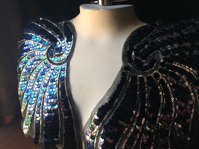 May include: Two black sequined fabric appliques with a blue and gold iridescent finish. The appliques are shaped like wings and are sewn onto a black mesh fabric.