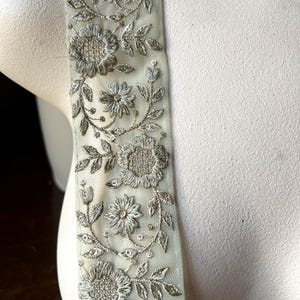 Może przedstawiać: Zbliżenie dekoracyjnego wykończenia z misternym haftem kwiatowym. Wykończenie jest w kolorze bladego zielonego, ze srebrną nicią używaną do kwiatów i liści. Wzór jest szczegółowy i elegancki, odpowiedni do ozdabiania odzieży lub rękodzieła.