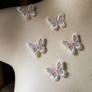 Puede incluir: Cinco delicados apliques de mariposa. Cada mariposa presenta un contorno bordado blanco con acentos rosas y dorados. Las mariposas están dispuestas sobre una superficie beige claro, creando un efecto caprichoso y decorativo.