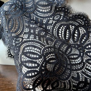 May include: Close-up of black lace fabric with intricate floral patterns. The lace is detailed with various shapes and textures, creating a delicate and elegant design. The fabric is draped over a mannequin, showcasing the lace's texture and drape.