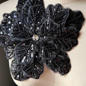 May include: A large, black sequined flower applique with a clear rhinestone center. The petals are outlined with a delicate black mesh. The applique is likely for embellishing clothing or accessories.