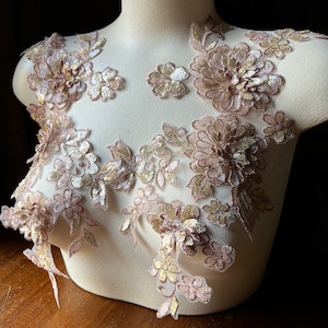 May include: A delicate floral lace applique in a blush pink and gold colour scheme. The lace features intricate floral designs with sequins and beads.
