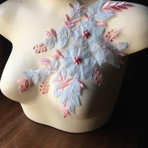 Może przedstawiać: Kwiatowa aplikacja z jasno-niebieskimi i różowymi haftowanymi detalami. Aplikacja zawiera delikatne kwiaty, liście i akcenty z koralików. Projekt jest prezentowany na manekinie.