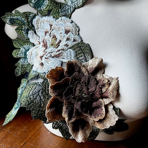 Pode incluir: Um par de apliques florais bordados em tons de marrom, branco e verde. Os apliques apresentam duas flores grandes com costuras detalhadas e acentos de folhas.