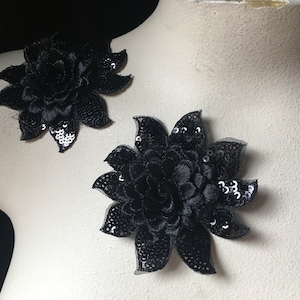 May include: Two black floral appliques with sequin accents. The appliques are made of fabric and feature a layered petal design. The sequins add a touch of sparkle to the flowers. The appliques are displayed on a mannequin.