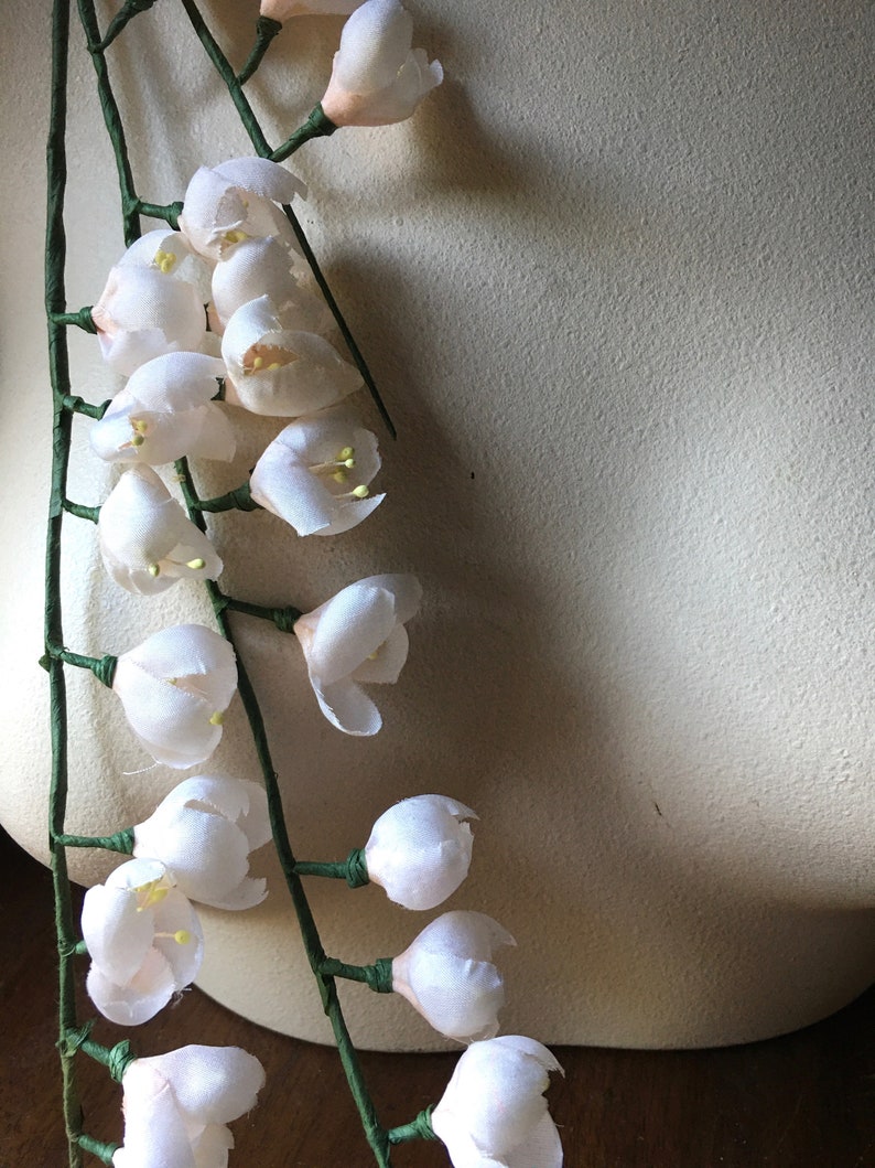 May include: A garland of delicate white silk flowers with green stems. The flowers are arranged in a cascading pattern, creating a romantic and elegant look.