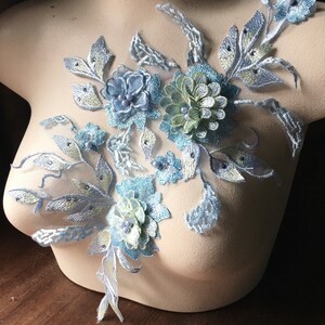 May include: A light blue and pale green floral lace applique featuring embroidered petals, leaves, and small round beads. The design is arranged in a sprawling, asymmetrical pattern on a sheer mesh backing, showcasing intricate stitching and layered textures.