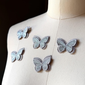 May include: Five silver butterfly-shaped embroidered patches arranged on a beige fabric dress form. Each patch features a detailed wing pattern with a metallic sheen and a stitched border, creating a textured appearance across the surface.