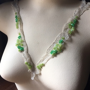 May include: A white lace trim with green bead embellishments. The trim is displayed on a white mannequin bust.