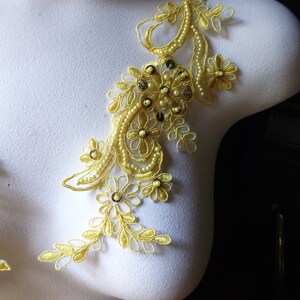 May include: A golden-yellow lace applique with floral and leaf designs, adorned with small pearls and sequins. The applique is displayed on a white mannequin, showcasing its intricate details and delicate craftsmanship. Ideal for embellishing garments.