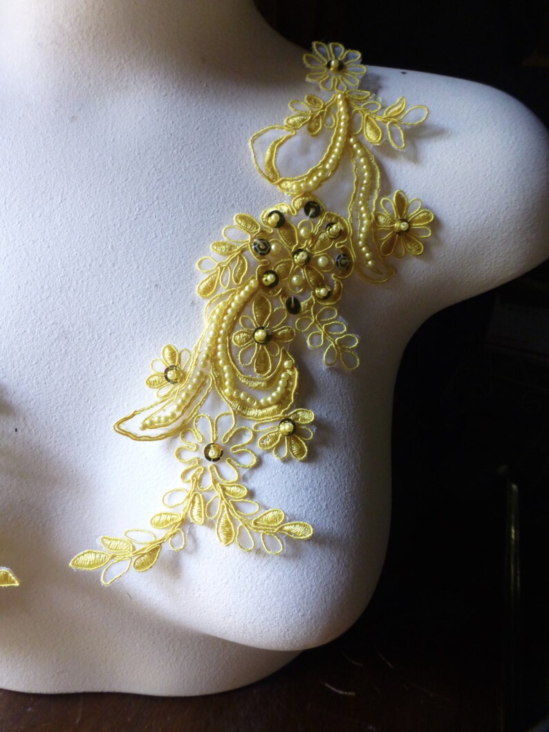 May include: A golden-yellow lace applique with floral and leaf designs, adorned with small pearls and sequins. The applique is displayed on a white mannequin, showcasing its intricate details and delicate craftsmanship. Ideal for embellishing garments.