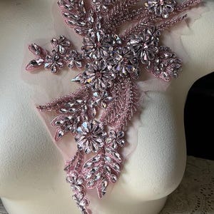 Puede incluir: Una aplicaci&oacute;n de cuentas y lentejuelas rosas con diamantes de imitaci&oacute;n transparentes en un dise&ntilde;o floral. La aplicaci&oacute;n est&aacute; sobre un fondo de tela transparente.
