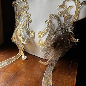 May include: Two gold sequined fabric appliques with a floral design. The appliques are on a white mannequin form and a wooden surface.