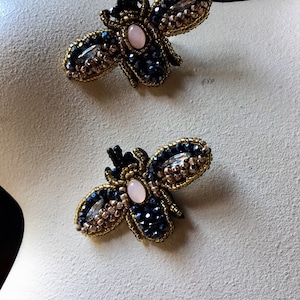 May include: Two handmade bee brooches. Each brooch features a body of dark blue crystals, gold-colored beaded wings, and a light pink oval stone. The bees are displayed on a white surface.