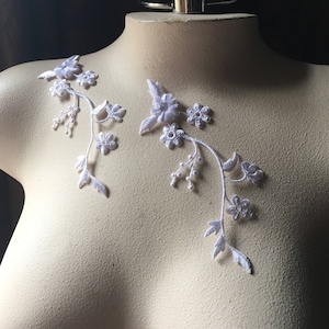 May include: Two white lace appliques with floral designs. Each applique features a delicate vine with small white flowers and leaves.