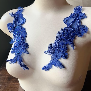 May include: Two pieces of royal blue lace applique, adorned with floral and beaded details, are displayed on a white mannequin. The lace is intricately designed with a delicate, flowing pattern, perfect for embellishing garments or crafts.