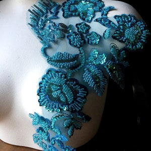 May include: Teal blue beaded floral applique with sequins. The applique is on a white background and is ideal for adding a touch of sparkle to any project.
