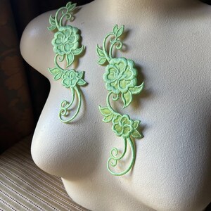 May include: Two green floral appliques with a delicate, intricate design. The appliques feature a repeating pattern of flowers and leaves, perfect for adding a touch of elegance to clothing or crafts.