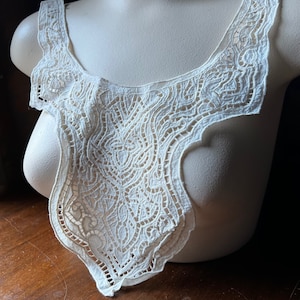 May include: Ivory-colored lace collar with intricate floral and geometric patterns. The collar is displayed on a mannequin, showcasing its delicate openwork design. The vintage-style collar is a decorative garment.